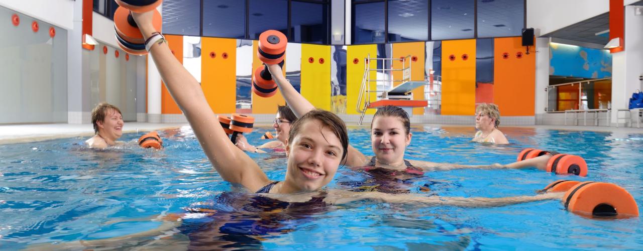 Mehrere Personen machen Aquagymnastik im Schwimmbecken mit orangen Schwimmbrettern und Hanteln, helle Schwimmhalle mit orangen und gelben Wänden