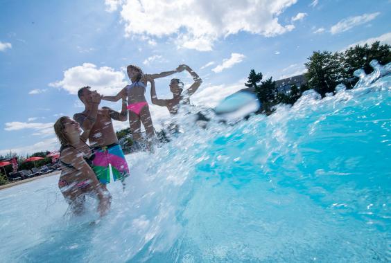 Vier Personen im Schwimmbad heben ein Mädchen in pinker Badehose in die Luft, Wasser spritzt, sonniger Himmel
