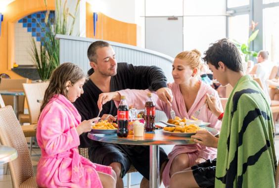 Vier Personen in Bademänteln sitzen an einem Tisch und essen Pommes mit Getränken in einem hellen Innenraum